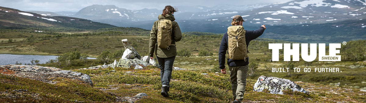 Two hikers wearing THULE outdoor backpacks and vests traverse a rugged mountain landscape with snowy peaks in the background. The THULE logo is visible on the right side of the image.