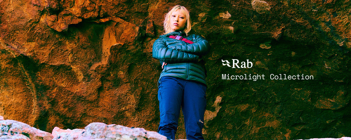 Person wearing a Rab Microlight insulated jacket and hiking trousers against a rocky cliff backdrop, with “Rab Microlight Collection” promotional text and Rab branding.