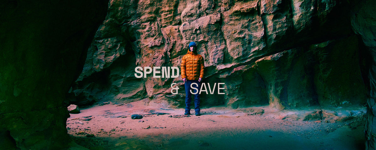 A person in an orange puffer jacket and blue pants stands inside a rocky cave or canyon, with large rock walls behind them and overlaid text reading 'SPEND & SAVE'.